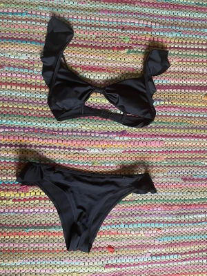 Svart bikini med volangdetaljer - Snygg svart bikini med volangdetaljer på både överdel och underdel. Överdelen har en dekorativ knytning framtill och justerbara axelband. Klassisk bikiniunderdel med matchande volanger. Perfekt för stranden eller poolen. Använd en gång å tvättad efteråt