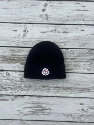 Svart Moncler mössa med logga - Snygg svart ribbstickad mössa från Moncler med uppvikt kant och klassisk Moncler-logga framtill. Perfekt accessoar för kalla dagar och enkel att matcha med olika outfits. Materialet känns mjukt och värmande.