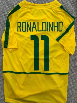 Gul Brasil Ronaldinho fotbollströja - Säljer en gul Brasilien fotbollströja med gröna detaljer och texten 'RONALDINHO' samt nummer 11 på ryggen. Tröjan har korta ärmar och är tillverkad i ett lätt och ventilerande material, perfekt för fotboll eller träning.