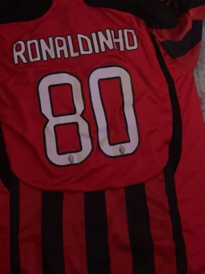 AC Milan Ronaldinho matchtröja - Säljer en klassisk AC Milan matchtröja från Adidas med Ronaldinho och nummer 80 på ryggen. Tröjan är röd och svart med vertikala ränder, klubbmärke på bröstet och Bwin som sponsor. Perfekt för dig som älskar fotboll och vill ha en ikonisk supportertröja.