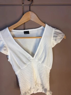 Vit smockad topp från Gina Tricot - Supersöt vit topp från Gina Tricot med smockad midja och volangkant nedtill. Toppen har korta puffärmar och djup v-ringning som ger en fräsch och somrig vibe. Perfekt att matcha med jeans eller kjol för en trendig look.