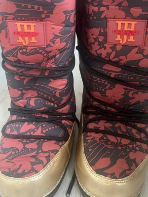 Ilse Jacobsen moon boots mönstrade - Moon boots från Ilse Jacobsen med svart och rött blommönster och detaljer i guld. Stora logotyper på både framsidan och hälen. Skorna har snörning och är vadderade för extra värme. Perfekta för kalla dagar när du vill sticka ut. Helt oanvända 
