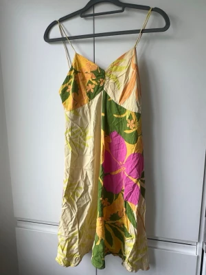 Blommig sommarklänning med smala band - Färgglad kortklänning med blommigt mönster i gult, grönt, rosa och beige. Klänningen har smala axelband och v-ringad urringning, perfekt för varma dagar. Tillverkad i ett lätt och luftigt material som passar bra till sommaren.