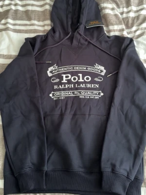Mörkblå hoodie från Polo Ralph Lauren - Snygg mörkblå hoodie från Polo Ralph Lauren med stor vit logga och text på bröstet. Klassisk huva med dragsko och ribbade muddar vid ärmar och nederkant. Tillverkad i mjukt bomullsmaterial för skön känsla. Perfekt för en avslappnad och trendig stil.