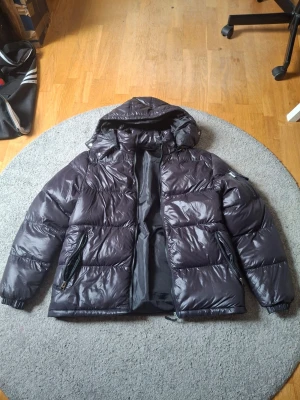 Svart pufferjacka från Brave Soul - Snygg svart pufferjacka från Brave Soul med glansig finish och stor huva. Jackan har dragkedja framtill, två sidofickor med dragkedja och breda quiltade partier. Perfekt för kalla dagar och riktigt varm tack vare det tjocka materialet.