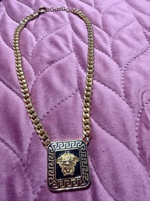 Guldigt halsband med lejonmotiv - Snyggt halsband i guldfärgad metall med en kraftig kedja och en fyrkantig svart berlock med ett detaljerat lejonhuvud i mitten. Runt berlocken finns ett klassiskt grekiskt mönster. Perfekt statement piece för dig som gillar unika accessoarer.