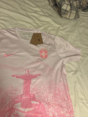 Rosa Brasil Nike fotbollströja - Säljer en snygg rosa fotbollströja från Nike med Brasil-logga och fem stjärnor på bröstet. Tröjan har motiv av Kristusstatyn och stadsmiljö i rosa nyanser. Materialet är Dri-FIT för optimal komfort och andningsförmåga. Perfekt för träning eller match.