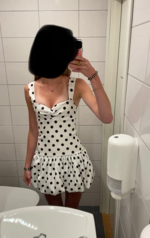 Vit prickig kort klänning - Supersöt vit kort klänning med svarta prickar och breda axelband. Klänningen har en volangig kjol och en snygg omlott-detalj vid bysten. Perfekt för dig som vill sticka ut med en lekfull och trendig look.