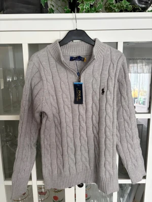 Grå stickad tröja Polo Ralph Lauren - Kabelstickad grå tröja från Polo Ralph Lauren med halv dragkedja och hög krage. Tröjan har ribbade muddar och den klassiska broderade loggan på bröstet. Perfekt för dig som gillar en stilren och tidlös look.