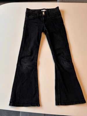 Svarta bootcut jeans med låg midja - Säljer ett par svarta bootcut jeans med låg midja och klassiska fem fickor. Jeansen har utsvängda ben och dubbla knappar i midjan. Snygg tvätt och skön passform, perfekta till sneakers eller boots.
