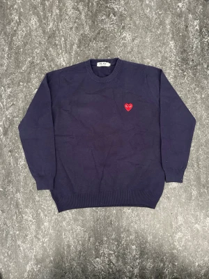 Mörkblå stickad tröja Comme des Garçons - (Priset kan diskuteras) Mörkblå stickad tröja från Comme des Garçons Play med det ikoniska röda hjärtat med ögon broderat på bröstet. Tröjan har rund halsringning, ribbade muddar och är tillverkad i mjukt material. Perfekt för dig som gillar streetwear och stilrena detaljer.