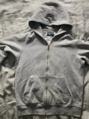 Grå zip-hoodie från Jack & Jones - Säljer en stilren grå hoodie från Jack & Jones med dragkedja och stora fickor framtill. Hoodien har en klassisk huva och är gjord i mjukt bomullsmaterial som är perfekt för chill dagar. Enkel och snygg design som funkar till det mesta.