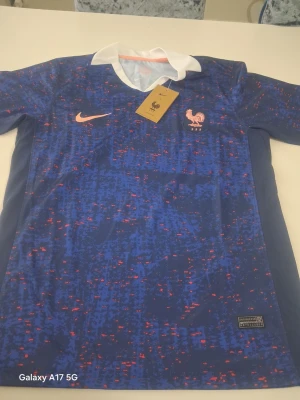 Blå Frankrike fotbollströja Nike - Säljer en blå fotbollströja från Nike med Frankrikes landslagsemblem och FFF-logga på bröstet. Tröjan har ett digitalt mönster i blått och rött, vit krage och korta ärmar. Perfekt för dig som älskar fotboll och vill stötta Les Bleus.