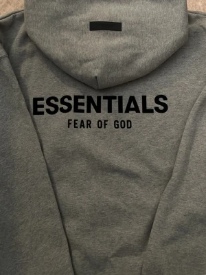Grå hoodie från Fear of God Essentials - Snygg grå hoodie från Fear of God Essentials med svart logga på bröstet och stor text på ryggen. Klassisk känguruficka framtill och huva. Tillverkad i mjukt bomullsmaterial som känns skönt mot huden. Perfekt för en avslappnad och trendig streetwear-look.