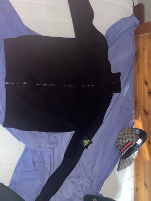 Svart stickad tröja från Stone Island - Säljer en svart stickad tröja från Stone Island med den ikoniska loggan på ärmen. Tröjan har ribbstickad struktur, rund hals och långa ärmar. Perfekt för dig som gillar streetwear och vill ha något stilrent och klassiskt.