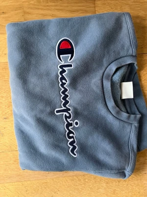 Blå sweatshirt från Champion - Snygg blå sweatshirt från Champion med det klassiska broderade logotypen i vitt, rött och blått på bröstet. Tröjan har rund halsringning och är tillverkad i mjukt bomullsmaterial. Perfekt för en avslappnad och sportig stil.