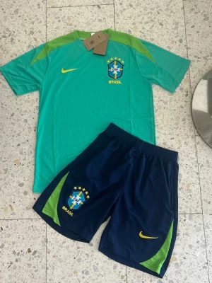 Brasilien fotbollsträningsset Nike - blå/grön/turkos - Snyggt träningsset från Nike med Brasilien-tema. Tröjan är turkos med gröna detaljer och har CBF-logga och gul Nike-swoosh. Perfekt nu inför sommar och VM☀️Pris kan alltid diskuteras vid snabb affär. Frakt inom 0-1 dagar!
