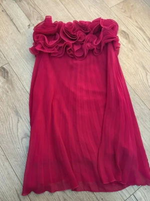  Ny  Trendig rosa  axelbandslös klänning från ZARA - Säljer en röd axelbandslös klänning från ZARA med volangdetaljer upptill och plisserad kjol. Klänningen har en elegant och romantisk look, perfekt för dig som vill sticka ut. Materialet är lätt och luftigt, vilket ger ett snyggt fall.