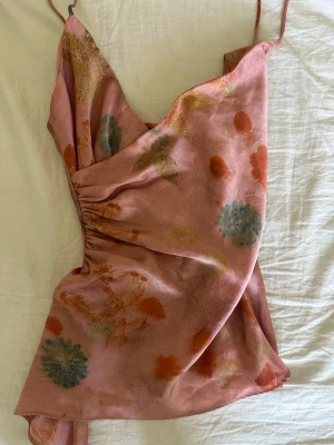 Blommig rosa linnetopp från Zara - Säljer en söt rosa linnetopp från Zara med tunna axelband och blommigt mönster i grönt, blått och orange. Toppen har en omlott-look och lätt glansigt material som känns lyxigt. Perfekt för dig som gillar unika detaljer och somriga vibes!