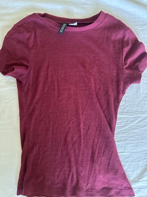 Vinröd t-shirt från Divided - En enkel vinröd t-shirt från Divided med rund halsringning och korta ärmar. Tillverkad i mjuk bomull som känns skön mot huden. Passformen är normal och färgen är djup och snygg, perfekt till jeans eller kjol. Har ett litet hål och är genomskinlig 