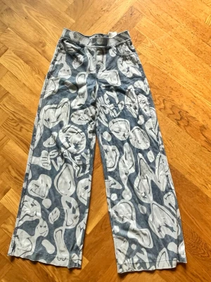 Blåa vida jeans med grafiskt mönster - Säljer ett par blåa jeans från H&M med vitt grafiskt mönster över hela byxan. Modellen är vid med hög midja och raka ben. Jeansen har en cool streetwear-vibe och är perfekta för dig som vill sticka ut lite extra.