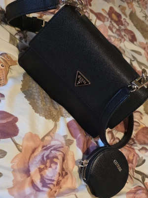 Svart handväska från Guess - Snygg svart handväska från Guess med strukturmönster och guldfärgade detaljer. Väskan har en justerbar axelrem och en matchande rund liten väska som hänger på sidan. Triangelformad Guess-logga framtill och ljus insida med fack. Köpt från Åhléns men sälja för att vill köpa nya