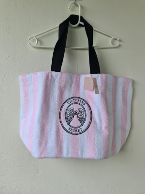 Randig shoppingväska Victoria's Secret - Säljer en stor shoppingväska från Victoria's Secret med rosa och vita ränder. Väskan har svarta handtag och ett broderat emblem med änglavingar och logga på framsidan. Materialet är mjukt, liknande frotté. Perfekt för att bära allt du behöver till stranden eller stan. Helt ny med lapparna kvar, pris disskuteras ej!