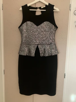 Svart klänning med silverpaljetter - Snygg svart klänning med silvriga paljetter över bysten och midjan. Klänningen har peplumdetalj och är ärmlös med rund halsringning. Perfekt för dig som vill sticka ut med lite extra glitter. Materialet känns stretchigt och bekvämt. Stolen M.
