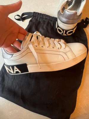 Dolce & Gabbana vita sneakers - Snygga vita sneakers från Dolce & Gabbana med silvriga detaljer på hälen och svart/vit logga baktill. Skorna har vita snören, diskreta röda sömmar och en chunky sula. Perfekta för dig som gillar stilrena men ändå lyxiga sneakers.