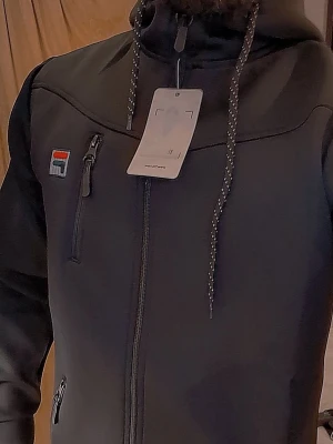 Svart hoodie från Fila med dragkedja - Snygg svart hoodie från Fila med hel dragkedja framtill och tre fickor med dragkedja. Klassisk Fila-logga på bröstet och ärmen. Huva med justerbara snören och sportig look. Tillverkad i mjukt och slitstarkt material, perfekt för en avslappnad stil. Helt ny fila hoodie s/m