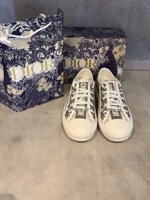 Dior Oblique låga sneakers - Säljer ett par snygga Dior Oblique sneakers i vitt och marinblått med klassiskt monogrammönster. Skorna har vita snören, gummisula och texten 'CHRISTIAN DIOR' på plösen. Perfekta för dig som vill ha en exklusiv och trendig look.