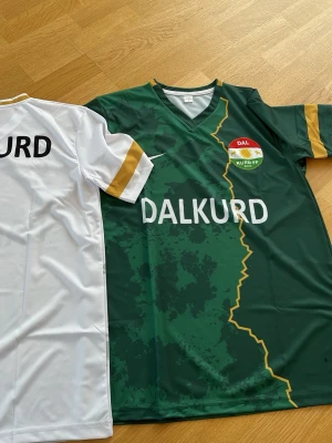 Dalkurd matchtröjor grön och vit - Två officiella Dalkurd matchtröjor, en grön med gul detalj och klubbmärke på bröstet samt en vit med svarta bokstäver och guldfärgade detaljer på ärmar och krage. Tillverkade i lätt och ventilerande funktionsmaterial, perfekta för fotbollsträning eller match. 500 FÖR BÅDA. FÖRPACKNING TILLKOMMER OCH DEN ÄR ALDRIG ANVÄND