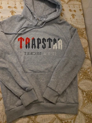 Grå hoodie från Trapstar - Snygg grå hoodie från Trapstar med broderad text i svart, vitt och rött på bröstet. Klassisk huva med dragsko och stor magficka framtill. Perfekt för en streetwear-look och riktigt skön att bära till vardags.