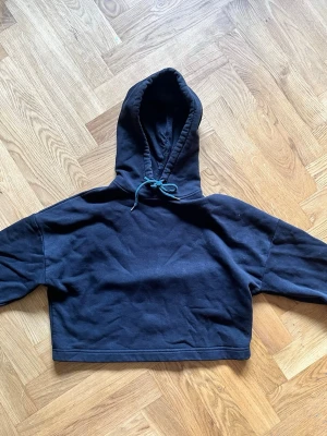 Svart cropped hoodie med huva - En svart cropped hoodie med huva och snörning framtill. Hoodien har långa ärmar. Materialet är mjukt och bekvämt, perfekt för en avslappnad och cool look. Passar dig som gillar streetstyle och enkel design.