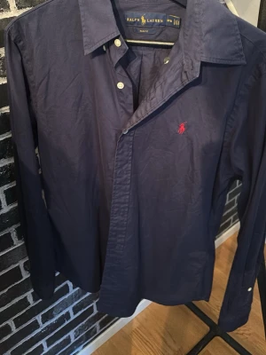 Marinblå skjorta från Ralph Lauren - Klassisk marinblå skjorta från Ralph Lauren med röd broderad logga på bröstet. Skjortan har långa ärmar, knappar framtill och en stilren krage. Skriv om frågor uppstår🤗