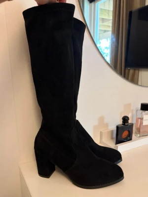 Svarta knähöga stövlar med klack - Snygga svarta knähöga stövlar i mockaimitation med bred klack och rundad tå. Perfekta för att lyfta vilken outfit som helst och ger en cool vibe. Stövlarna har en stilren design utan synliga detaljer eller dekorationer.