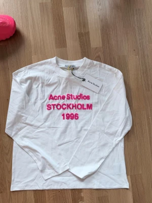 Vit långärmad topp Acne Studios - Vit långärmad topp från Acne Studios med rund hals och rosa broderad text 'Acne Studios STOCKHOLM 1996' framtill. Tillverkad i mjuk bomull och har en clean, minimalistisk look med en oversized känsla.