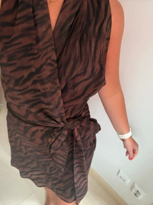 Brun omlottklänning med zebramönster - Säljer en kort brun omlottklänning med svart zebramönster. Klänningen har knyt i midjan och är ärmlös med en snygg v-ringning. Perfekt för dig som vill ha en trendig och unik look.