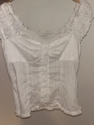 Vit broderad blus med volang - Söt vit blus med broderade detaljer och volangkant. Toppen har breda axelband, smock upptill och dekorativa små hålbroderier över hela tyget. Perfekt för en somrig look och ger romantiska vibbar.