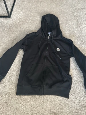 Svart hoodie från Moncler - Säljer en svart hoodie från Moncler med dragkedja framtill och klassisk Moncler-logga på bröstet. Hoodien har huva med dragsko, två stora fickor fram och ribbade muddar vid ärmslut och nederkant. Perfekt för en chill och stilren look. Nyligen köpt från deras hemsida, har tagg och påsen kvar.