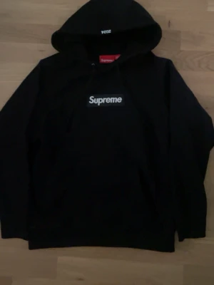 Svart Supreme hoodie - Svart hoodie från Supreme med klassisk vit Supreme-logga på bröstet. Tröjan har huva med dragsko och en röd Supreme-tag i nacken. Perfekt för dig som gillar streetwear och vill sticka ut med en ikonisk look.