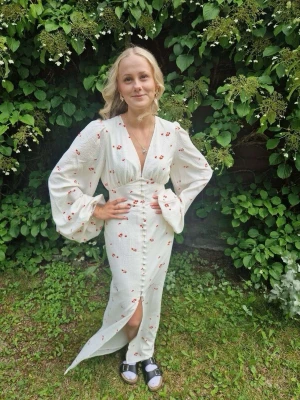 Vit långklänning med blommor - Vit långklänning med små röda blommor, v-ringad och med knappar framtill med vida och långa ärmar. Perfekt för sommarens alla tillfällen. Från Adoore. (Knapparna högst upp är lite ”uttöjda”) men fick klänningen av ett ex så det är inget fel på den så jag kommer bara inte använda den igen! Därför säljer jag den.