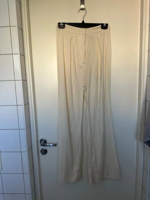 Vida beige med hög midja 36 - Snygga vida i beige med hög midja och pressveck framtill. Byxorna har en klassisk look med dragkedja och knappstängning. Perfekta för en avslappnad men stilren outfit. Materialet är lätt och luftigt linne. Några fläckar som nog enkelt går bort med fläckborttagningsmedel
