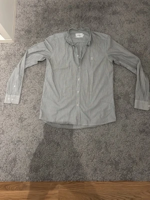 Blåvit randig skjorta från Les Deux - Snygg Blåvit skjorta från Les Deux med tunna ränder och klassisk krage. Skjortan har långa ärmar, vita knappar framtill och på manschetterna samt en liten broderad logga på bröstet. Perfekt till jeans eller chinos för en clean look.