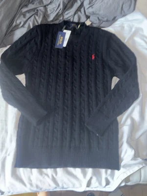 Svart kabelstickad tröja Polo Ralph Lauren - Kabelstickad svart tröja från Polo Ralph Lauren med klassisk röd logga broderad på bröstet. Tröjan har rund halsringning, långa ärmar och ribbade muddar. Perfekt för en stilren och tidlös look.
