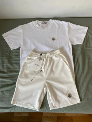 Vit Moncler t-shirt och shorts  - Klassisk vit t-shirt från Moncler med rund hals och korta ärmar. Tillverkad i mjuk bomull och prydd med Monclers ikoniska logotyp på bröstet. Perfekt basic-plagg för en clean och stilren look.
