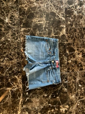 Blå jeansshorts med fransig kant - Säljer ett par klassiska blå jeansshorts med fransig kant nertill. Shortsen har fem fickor, bälteshällor och knappgylf. Perfekta för varma dagar och har en avslappnad vibe.