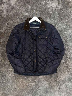 Mörkblå quiltad jacka från polo ralph lauren - Quiltad mörkblå jacka från polo ralph lauren med bruna detaljer på kragen och tryckknappar. Jackan har en bröstficka med knapp, två sidofickor och dragkedja framtill. Perfekt för kyligare dagar och har en klassisk, stilren look.