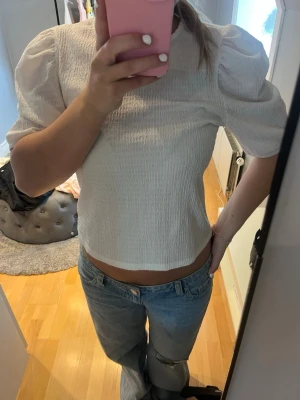 Vit puffärmad croppad blus - Säljer en vit croppad blus med puffärmar och lätt rynkad struktur. Toppen har rund halsringning och trekvartsärm, vilket ger en trendig och fräsch look. Perfekt att matcha med jeans eller kjol för en stilren outfit.
