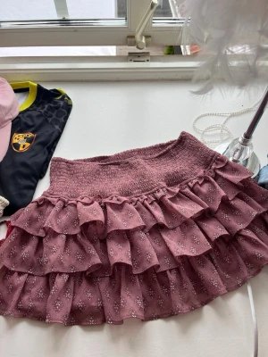 Rosa volangkjol med mönster - Supersöt kort volangkjol i dammigt rosa med små blommiga mönster. Kjolen har bred resår i midjan och flera lager volanger som ger en fluffig och lekfull look. Perfekt för dig som gillar romantisk och trendig stil.